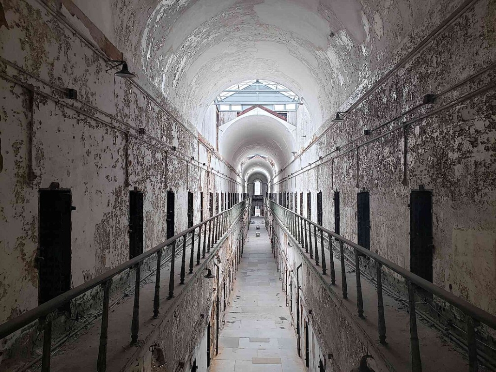 This photograph showcases a cellblock within the historic Eastern State Penitentiary. The image depicts a long, vaulted corridor flanked by rows of cell doors, illustrating the penitentiary's once strict regime of solitary confinement.