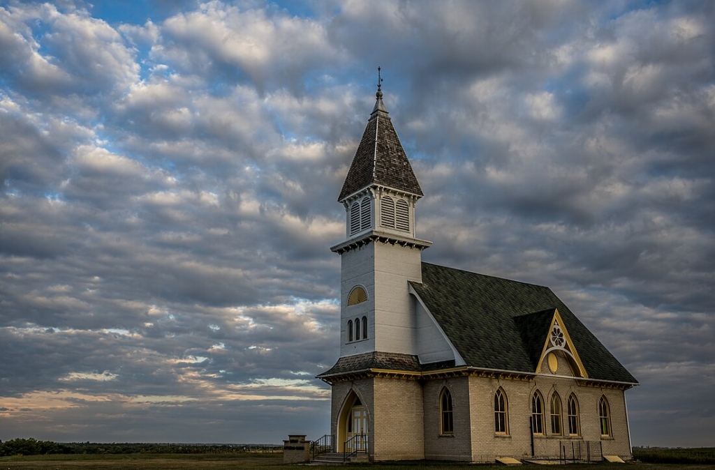 This photograph captures the Norway Lutheran Church and Cemetery, an exquisite example of Late Gothic Revival architecture, located near Denbigh, North Dakota. Built in 1907, the church stands against a dramatic sky, its white exterior and elegant steeple contrasting with the surrounding landscape.