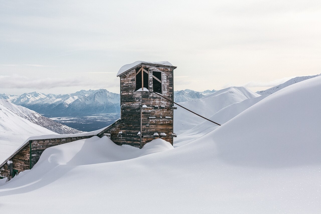 This image captures one of the historical structures associated with the Independence Mines, located in the Talkeetna Mountains of Alaska. The photograph, taken in 2018, shows the aged wooden structure partially enveloped by snow, with its weathered walls standing in stark contrast to the pristine white landscape.