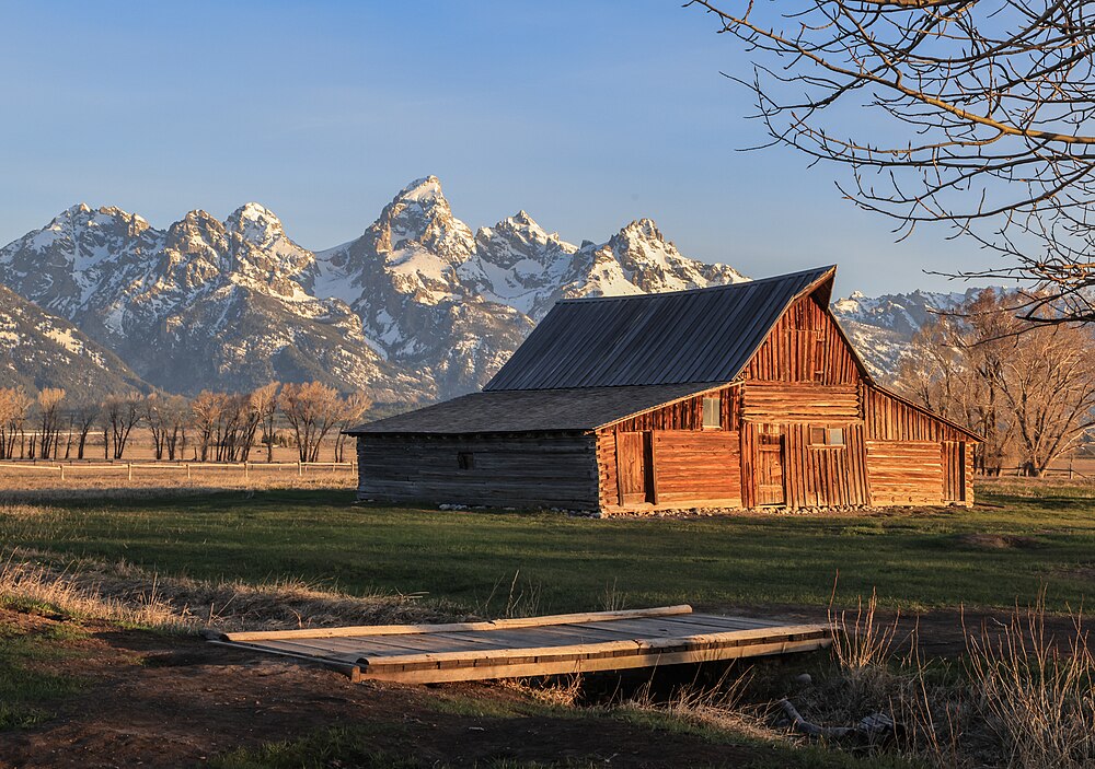 The photograph captures the warm glow of sunrise on one of the iconic Mormon Row Historic District barns with the Teton Range providing a stunning backdrop. This Historic District within Grand Teton National Park, Wyoming, is known for its scenic views and historic significance.