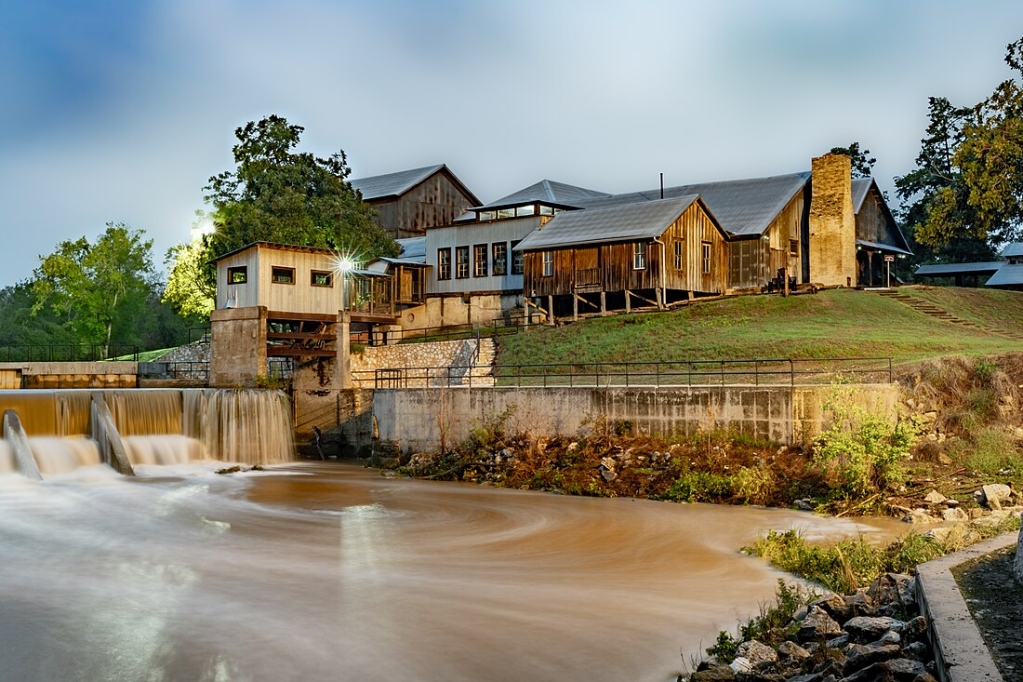 This photograph captures the serene twilight atmosphere at the Zedler Mill complex in Luling, Texas. The image is taken after a recent rainstorm, which has contributed to the increased flow and the muddy appearance of the water cascading over the spillway.